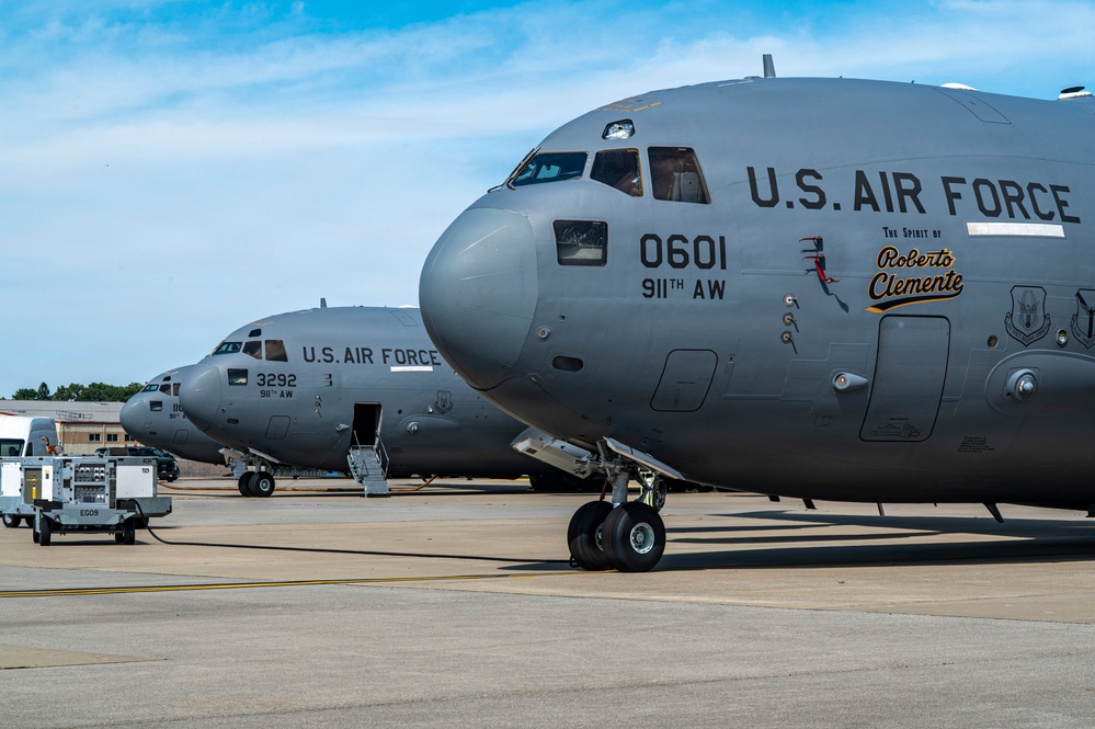 Two U.S. Air Force C-17 Globemaster III aircraft are parked on a flight line ramp, likely at a military air base. The foreground aircraft bears the tail number 0601, assigned to the 911th Airlift Wing, and features a special nose art dedication reading 'The Spirit of Roberto Clemente.' Ground support equipment and vehicles are visible between and around the aircraft.