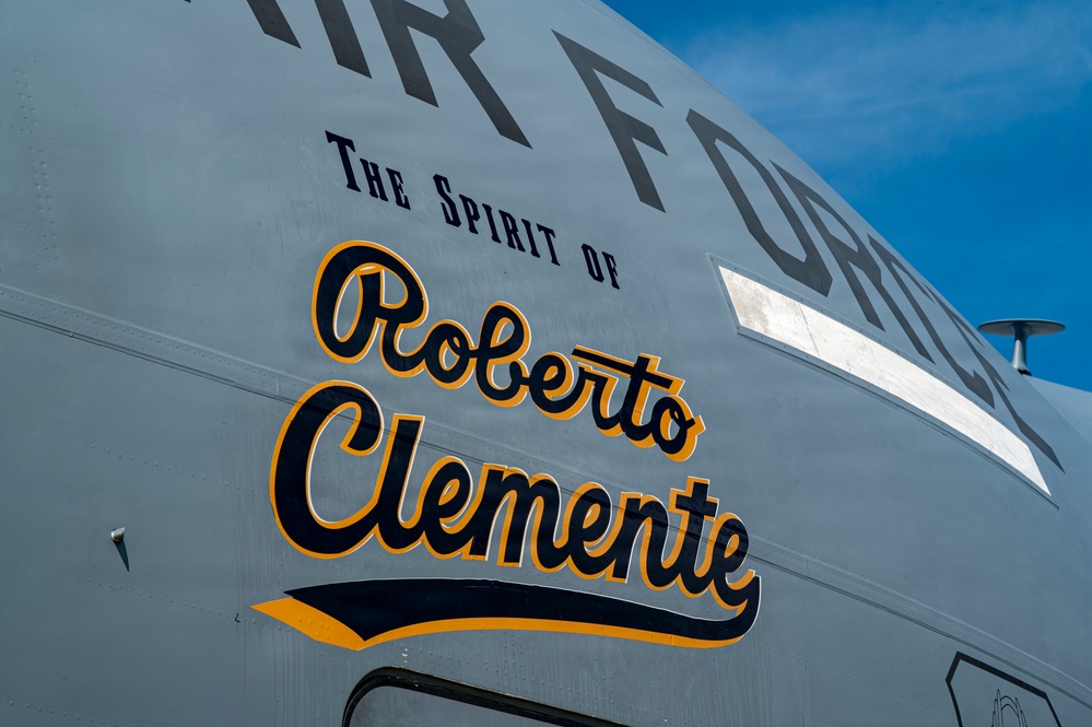 The image shows the fuselage of a U.S. Air Force aircraft named 'The Spirit of Roberto Clemente.' The name is painted in a stylized script font using black and gold/yellow colors, reminiscent of Pittsburgh Pirates baseball team colors. The aircraft appears to be a large military transport or tanker plane photographed against a blue sky.
