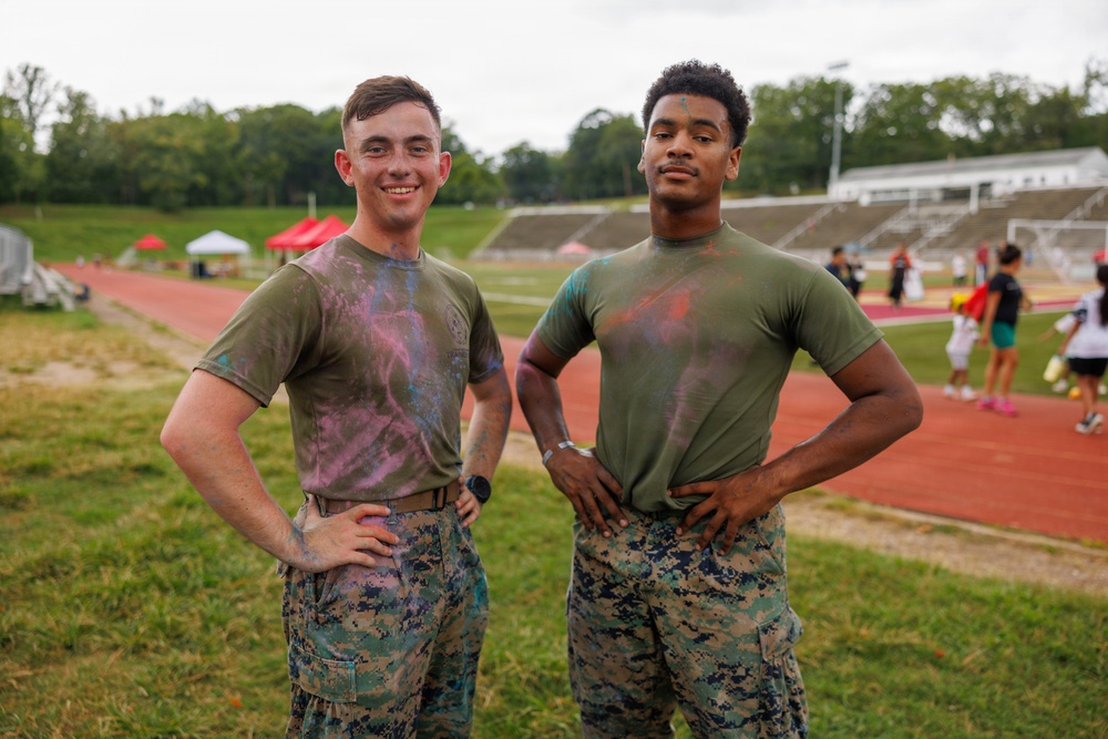 Two individuals in U.S. Marine Corps uniforms (olive drab t-shirts and MARPAT digital camouflage trousers) pose together at what appears to be a color run or Holi-style event at a military installation track and field facility. Both are covered in colored powder residue, suggesting participation in a recreational or morale event. They are standing on a grass infield beside a red running track with event tents and other participants visible in the background.