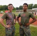 Two individuals in U.S. Marine Corps uniforms (olive drab t-shirts and MARPAT digital camouflage trousers) pose together at what appears to be a color run or Holi-style event at a military installation track and field facility. Both are covered in colored powder residue, suggesting participation in a recreational or morale event. They are standing on a grass infield beside a red running track with event tents and other participants visible in the background.