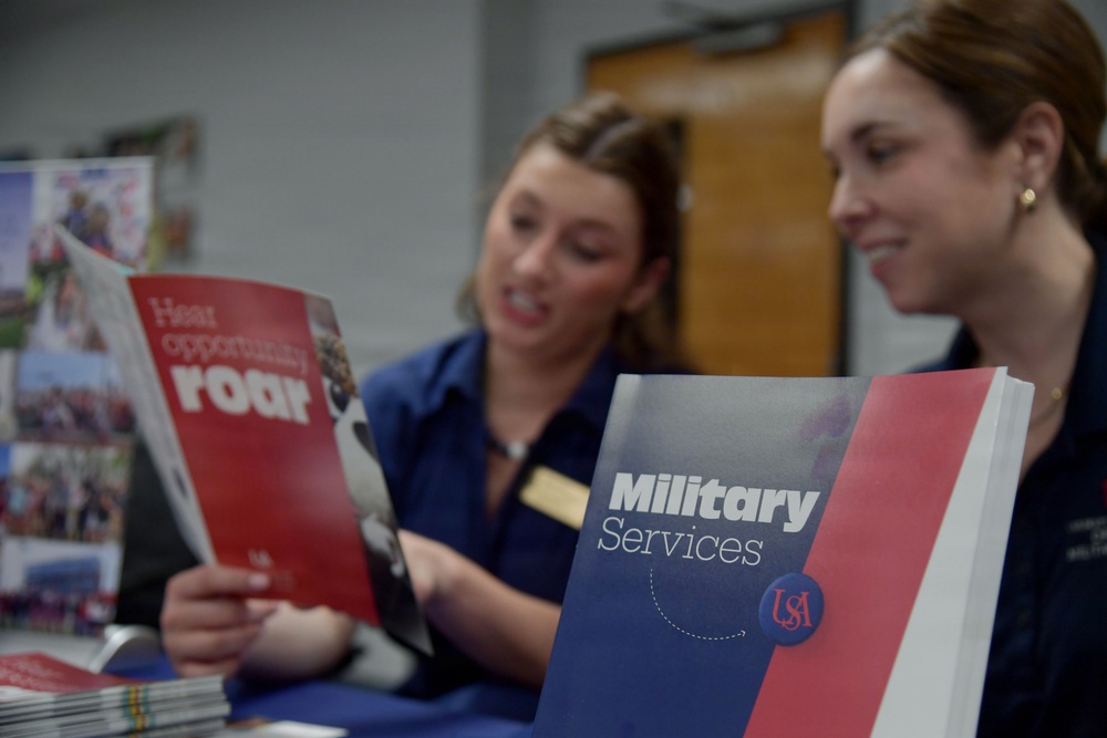Two women in matching navy blue uniforms or shirts are at what appears to be an informational or recruitment table. One woman holds a red brochure reading 'Hear opportunity roar' while a 'Military Services USA' brochure is prominently displayed in the foreground. The setting appears to be an educational or outreach event, likely representing the University of South Alabama (USA).