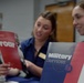 Two women in matching navy blue uniforms or shirts are at what appears to be an informational or recruitment table. One woman holds a red brochure reading 'Hear opportunity roar' while a 'Military Services USA' brochure is prominently displayed in the foreground. The setting appears to be an educational or outreach event, likely representing the University of South Alabama (USA).
