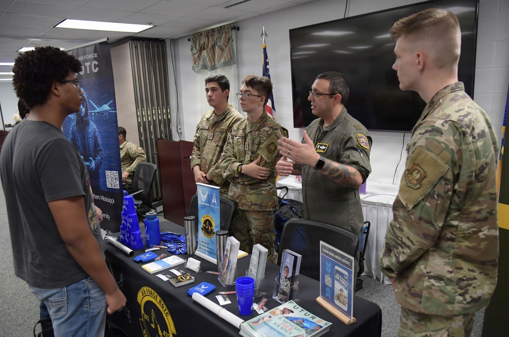 A military recruitment or information event featuring Air Force ROTC personnel speaking with civilians and other uniformed individuals. Three individuals in military uniforms (camouflage and flight suit) are engaged in conversation at a display table with a young civilian. The table is covered with Air Force ROTC promotional materials, brochures, and branded merchandise.