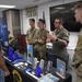 A military recruitment or information event featuring Air Force ROTC personnel speaking with civilians and other uniformed individuals. Three individuals in military uniforms (camouflage and flight suit) are engaged in conversation at a display table with a young civilian. The table is covered with Air Force ROTC promotional materials, brochures, and branded merchandise.