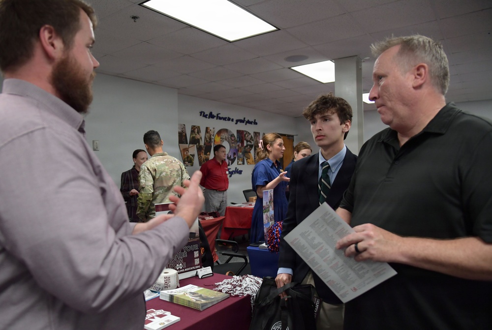 The image depicts what appears to be a college or career fair taking place in an indoor venue. Multiple booths are set up with representatives speaking to attendees, including a young man in a suit and tie accompanied by an older man holding informational materials. A wall mural with the word 'WORK' is visible in the background along with various exhibitor tables.
