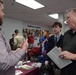 The image depicts what appears to be a college or career fair taking place in an indoor venue. Multiple booths are set up with representatives speaking to attendees, including a young man in a suit and tie accompanied by an older man holding informational materials. A wall mural with the word 'WORK' is visible in the background along with various exhibitor tables.