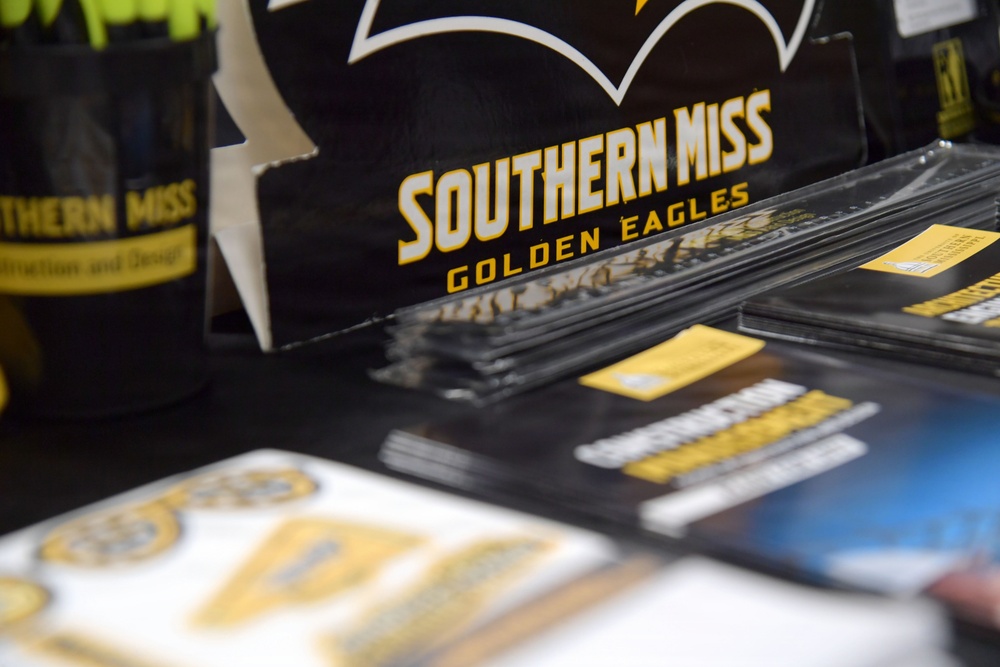 The image shows a promotional display table for the University of Southern Mississippi, featuring branded merchandise and informational materials. The table is covered with black tablecloth and displays various items in the school's black and gold colors. This appears to be a recruitment fair, college expo, or university event booth.