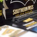 The image shows a promotional display table for the University of Southern Mississippi, featuring branded merchandise and informational materials. The table is covered with black tablecloth and displays various items in the school's black and gold colors. This appears to be a recruitment fair, college expo, or university event booth.
