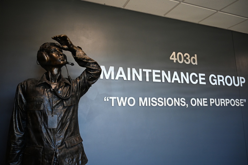 The image shows a bronze statue of a military maintenance technician wearing a flight suit, headset, and dog tags, positioned in front of a dark blue wall. The wall displays signage for the '403d Maintenance Group' with the motto 'Two Missions, One Purpose.' The statue depicts the figure shielding their eyes and looking upward, likely simulating aircraft observation.