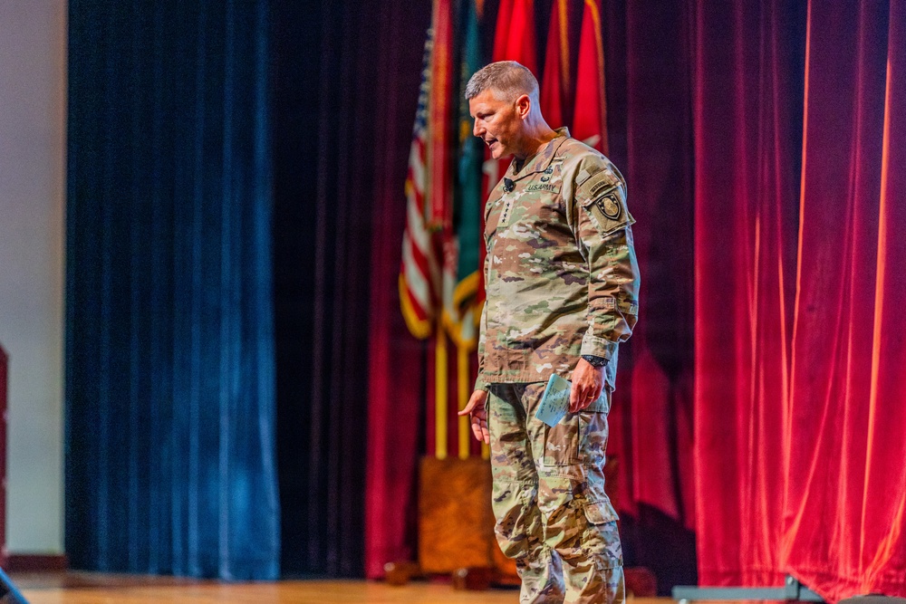 A U.S. Army officer in Operational Camouflage Pattern (OCP) uniform is standing on a stage, appearing to speak or present. He is holding what appears to be a small notecard or booklet and is looking downward. The stage is decorated with red curtains and multiple flags including the U.S. flag and what appear to be military unit colors.