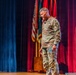 A U.S. Army officer in Operational Camouflage Pattern (OCP) uniform is standing on a stage, appearing to speak or present. He is holding what appears to be a small notecard or booklet and is looking downward. The stage is decorated with red curtains and multiple flags including the U.S. flag and what appear to be military unit colors.