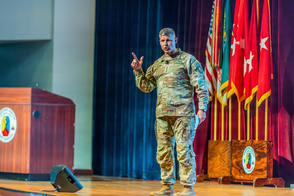 A U.S. Army general officer in OCP (Operational Camouflage Pattern) uniform is delivering a presentation on a stage, gesturing with one raised finger. He stands before a backdrop of military flags and a wooden podium bearing an insignia. The setting appears to be a formal military auditorium or conference hall.