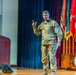 A U.S. Army general officer in OCP (Operational Camouflage Pattern) uniform is delivering a presentation on a stage, gesturing with one raised finger. He stands before a backdrop of military flags and a wooden podium bearing an insignia. The setting appears to be a formal military auditorium or conference hall.