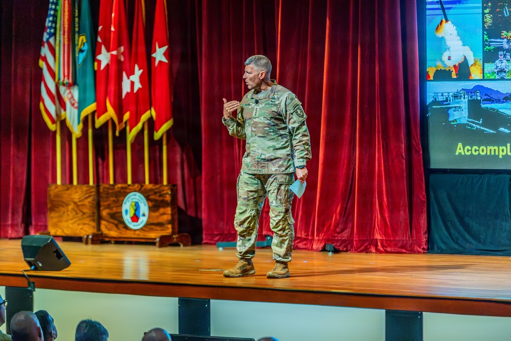 A U.S. Army officer in camouflage uniform is delivering a presentation on a wooden stage before an audience. Behind him are multiple military flags including the U.S. flag and Army unit colors, along with a projection screen displaying what appears to be military equipment imagery and the partial word 'Accompl'. A wooden podium with a military unit seal is visible to the left.
