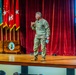 A U.S. Army officer in camouflage uniform is delivering a presentation on a wooden stage before an audience. Behind him are multiple military flags including the U.S. flag and Army unit colors, along with a projection screen displaying what appears to be military equipment imagery and the partial word 'Accompl'. A wooden podium with a military unit seal is visible to the left.