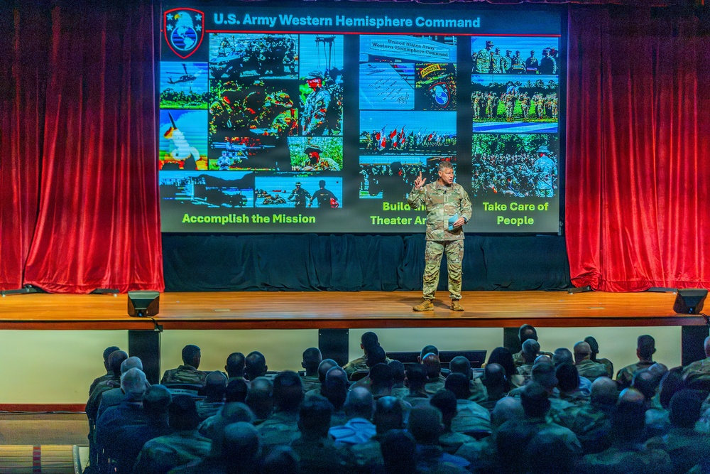 A U.S. Army soldier in military uniform is presenting on stage to a large audience of military personnel. The presentation slide behind them displays 'U.S. Army Western Hemisphere Command' with multiple military operation photos and three key pillars: 'Accomplish the Mission,' 'Build [Theater Army],' and 'Take Care of People.' The venue appears to be a formal auditorium with red curtains framing the stage.