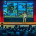 A U.S. Army soldier in military uniform is presenting on stage to a large audience of military personnel. The presentation slide behind them displays 'U.S. Army Western Hemisphere Command' with multiple military operation photos and three key pillars: 'Accomplish the Mission,' 'Build [Theater Army],' and 'Take Care of People.' The venue appears to be a formal auditorium with red curtains framing the stage.