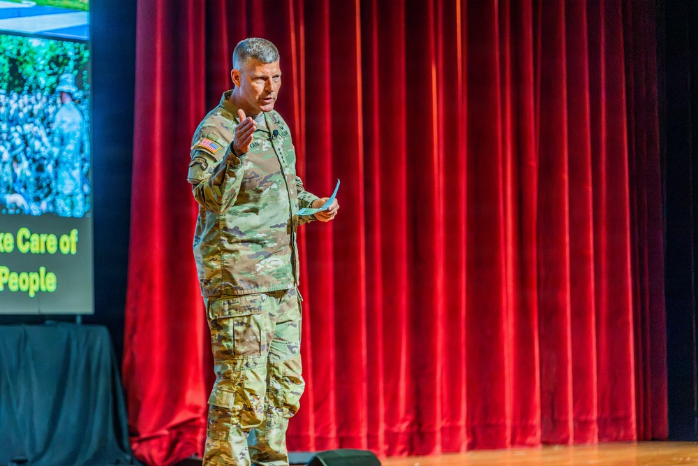 A U.S. Army soldier in MultiCam/OCP camouflage uniform is delivering a presentation on a stage. He is gesturing with one hand while holding what appears to be note cards in the other, standing in front of a red velvet curtain backdrop. A projection screen to his left displays text partially reading 'ke Care of People' alongside military imagery.