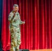 A U.S. Army soldier in MultiCam/OCP camouflage uniform is delivering a presentation on a stage. He is gesturing with one hand while holding what appears to be note cards in the other, standing in front of a red velvet curtain backdrop. A projection screen to his left displays text partially reading 'ke Care of People' alongside military imagery.