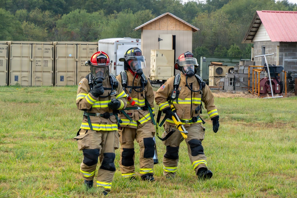Three firefighters in full personal protective equipment (PPE) walk across a grassy field, likely during a training exercise. They are wearing tan turnout gear with yellow reflective striping, SCBA (self-contained breathing apparatus) masks, and helmets numbered '188'. The background includes shipping containers marked 'USFU', training structures, and wooded terrain consistent with a fire training facility.