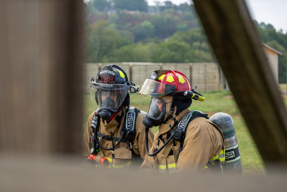 Two firefighters in full personal protective equipment are photographed through a gap in a wooden structure, suggesting a training exercise or active scene. Both wear MSA brand self-contained breathing apparatus (SCBA) and turnout gear. One wears a black helmet marked '188' and 'FIREFIGHTER,' while the other wears a red helmet.