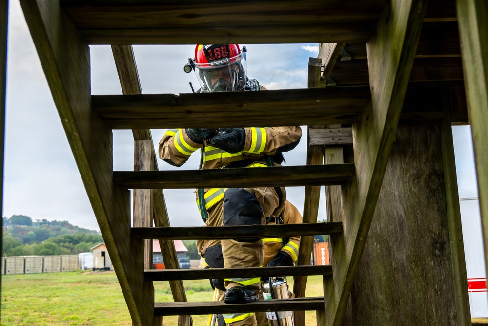 A firefighter in full personal protective equipment is climbing a wooden ladder structure, viewed from below at an upward angle. The individual is wearing a red helmet marked with the number '182', a full-face respirator/SCBA mask, tan turnout gear with yellow reflective striping, and black gloves. The setting appears to be an outdoor training facility based on the wooden tower structure and open field visible in the background.