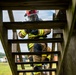 A firefighter in full personal protective equipment is climbing a wooden ladder structure, viewed from below at an upward angle. The individual is wearing a red helmet marked with the number '182', a full-face respirator/SCBA mask, tan turnout gear with yellow reflective striping, and black gloves. The setting appears to be an outdoor training facility based on the wooden tower structure and open field visible in the background.