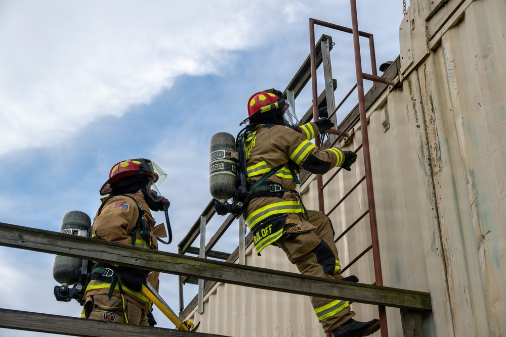 Two firefighters in full turnout gear are climbing a metal ladder on the exterior of a training structure. Both are equipped with SCBA (Self-Contained Breathing Apparatus) tanks and wearing red helmets with face shields. The scene appears to be a firefighter training exercise at an outdoor facility.