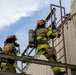 Two firefighters in full turnout gear are climbing a metal ladder on the exterior of a training structure. Both are equipped with SCBA (Self-Contained Breathing Apparatus) tanks and wearing red helmets with face shields. The scene appears to be a firefighter training exercise at an outdoor facility.