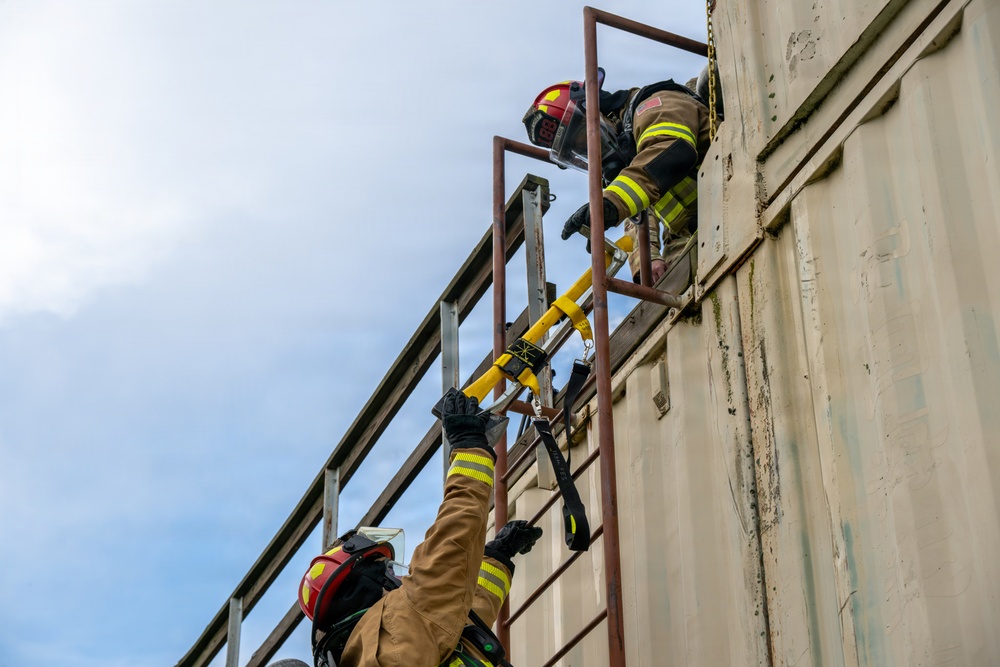 Two firefighters in full turnout gear and red helmets are climbing a ladder on the exterior of a large metal structure, likely a shipping container or industrial building. The image is taken from a low angle looking upward, showing one firefighter near the top and another ascending below. A yellow tool or piece of equipment is secured to the ladder railing, suggesting this is a training exercise or active rescue operation.