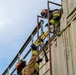 Two firefighters in full turnout gear and red helmets are climbing a ladder on the exterior of a large metal structure, likely a shipping container or industrial building. The image is taken from a low angle looking upward, showing one firefighter near the top and another ascending below. A yellow tool or piece of equipment is secured to the ladder railing, suggesting this is a training exercise or active rescue operation.