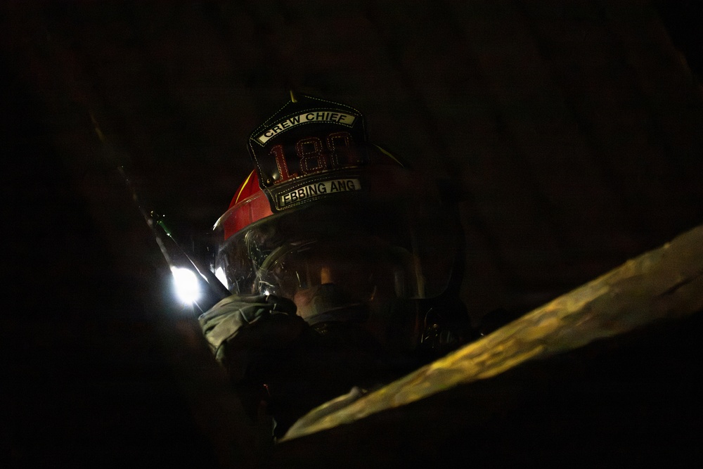 A firefighter wearing a red helmet marked 'CREW CHIEF' with the number '182' and name 'EBBING ANG' is photographed in very low-light conditions. The individual is equipped with a full face shield and appears to be operating in a dark, confined or debris-filled environment. A bright light source is visible to the left, likely a helmet-mounted or handheld flashlight.