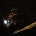 A firefighter wearing a red helmet marked 'CREW CHIEF' with the number '182' and name 'EBBING ANG' is photographed in very low-light conditions. The individual is equipped with a full face shield and appears to be operating in a dark, confined or debris-filled environment. A bright light source is visible to the left, likely a helmet-mounted or handheld flashlight.