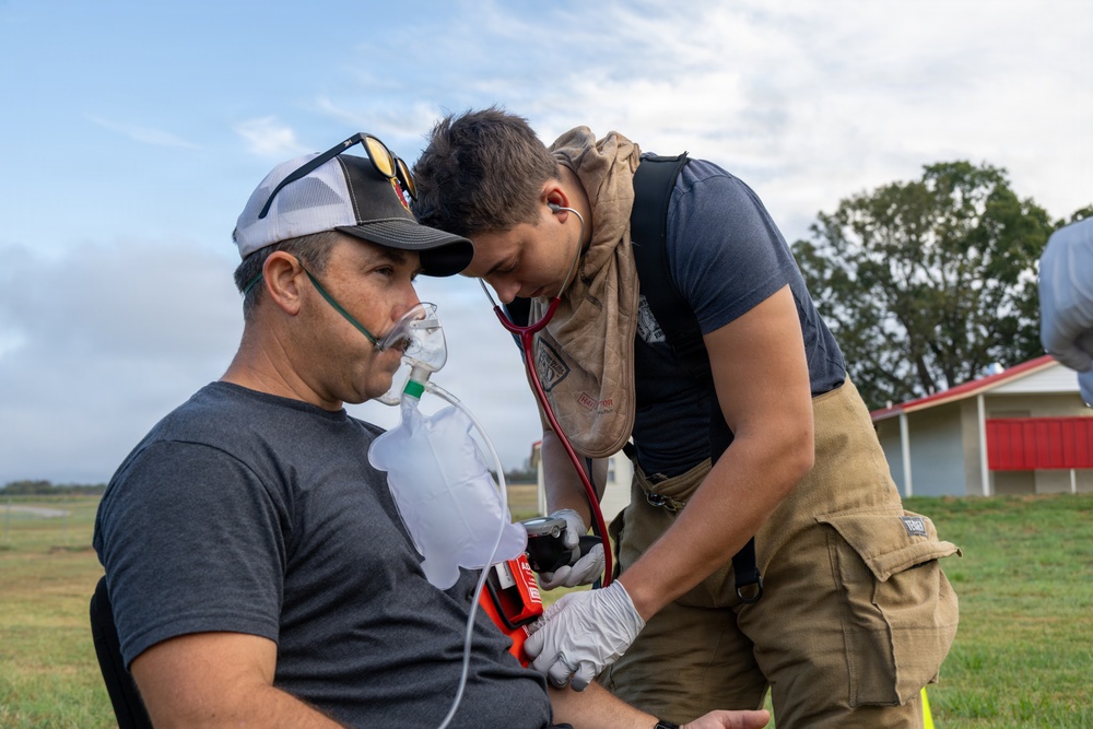 A first responder or firefighter in partial turnout gear is providing medical assistance to a seated male patient outdoors. The patient is wearing an oxygen mask with a reservoir bag while the responder uses a stethoscope and appears to be taking a blood pressure reading. The scene appears to be in a rural or open field setting, possibly during an emergency response or training exercise.