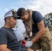 A first responder or firefighter in partial turnout gear is providing medical assistance to a seated male patient outdoors. The patient is wearing an oxygen mask with a reservoir bag while the responder uses a stethoscope and appears to be taking a blood pressure reading. The scene appears to be in a rural or open field setting, possibly during an emergency response or training exercise.