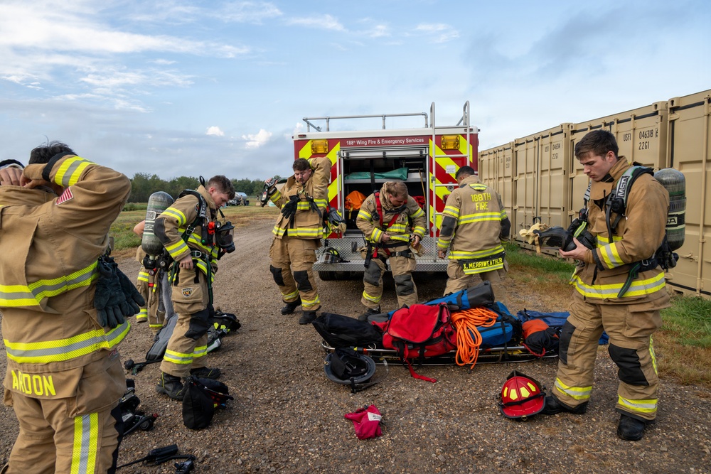 A group of firefighters from the 188th Wing Fire &amp; Emergency Services are gearing up or staging equipment near a red fire truck on a gravel surface. Multiple personnel in tan/yellow turnout gear with reflective striping are donning SCBA (self-contained breathing apparatus) equipment and organizing rescue gear. The scene appears to be a training exercise or pre-incident staging area, with shipping containers visible in the background.