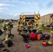 A group of firefighters from the 188th Wing Fire &amp; Emergency Services are gearing up or staging equipment near a red fire truck on a gravel surface. Multiple personnel in tan/yellow turnout gear with reflective striping are donning SCBA (self-contained breathing apparatus) equipment and organizing rescue gear. The scene appears to be a training exercise or pre-incident staging area, with shipping containers visible in the background.
