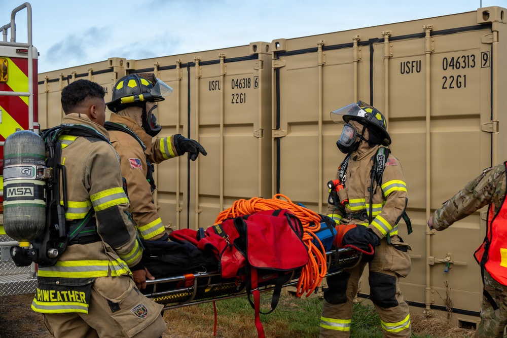 Three firefighters in full turnout gear and SCBA (Self-Contained Breathing Apparatus) are carrying a stretcher loaded with rescue equipment near tan military-style shipping containers. One firefighter appears to be giving directions while the others transport the gurney. The scene appears to be a training exercise or emergency response drill, likely at a military installation.
