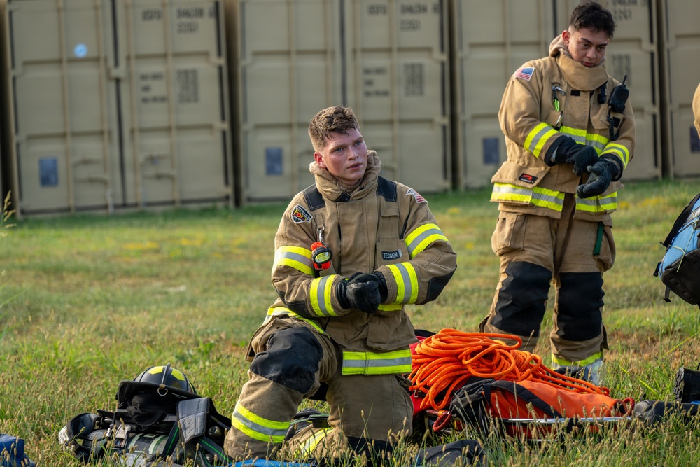 Two firefighters in full turnout gear are positioned in a grassy field near large storage containers. The foreground firefighter is kneeling and appears to be adjusting or donning gloves, while the background firefighter stands and also works with gloves. Firefighting equipment including an orange rope/hose coil, helmets, and bags are laid out on the ground nearby.