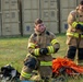 Two firefighters in full turnout gear are positioned in a grassy field near large storage containers. The foreground firefighter is kneeling and appears to be adjusting or donning gloves, while the background firefighter stands and also works with gloves. Firefighting equipment including an orange rope/hose coil, helmets, and bags are laid out on the ground nearby.