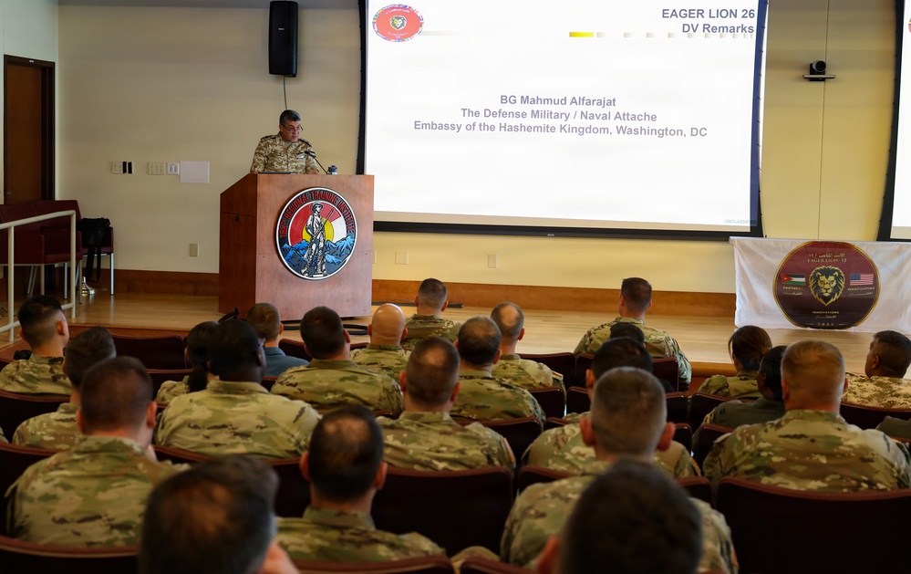 A military briefing or ceremony is taking place in an auditorium-style room where a uniformed officer is speaking at a podium. The presentation slide identifies the speaker as BG Mahmud Alfarajat, the Defense Military/Naval Attaché from the Embassy of the Hashemite Kingdom in Washington, DC, for the 'Eager Lion 26' exercise. An audience of military personnel in camouflage uniforms is seated facing the speaker.