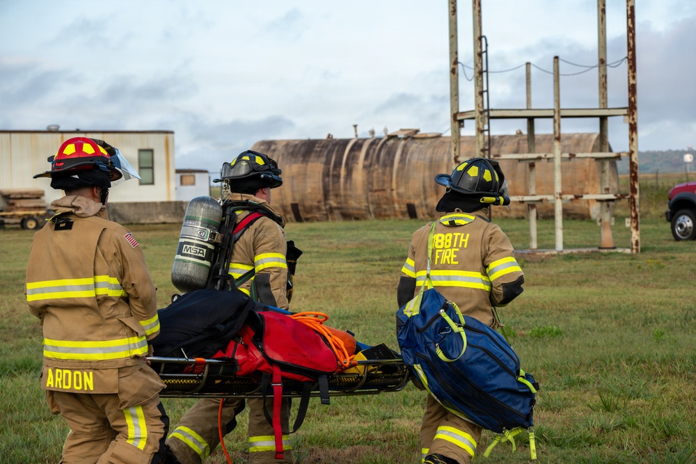Three firefighters in full turnout gear are carrying a stretcher/backboard with emergency equipment across a grassy field toward what appears to be a training facility with large industrial tanks. The personnel are identifiable by unit markings, with one labeled 'ARDON' and another marked '188TH FIRE'. This appears to be a firefighting training exercise or emergency response drill.