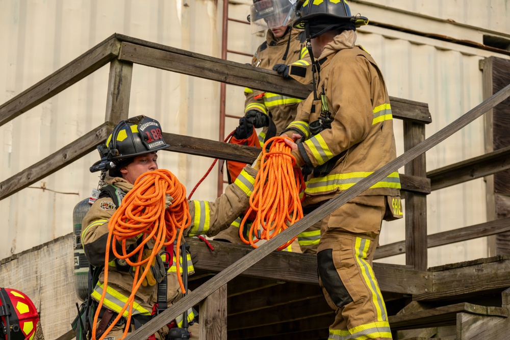 Three firefighters in full turnout gear are ascending a wooden staircase at what appears to be a training facility. They are passing and carrying coiled orange rescue ropes as they climb the stairs. The scene depicts a coordinated training exercise involving rope handling and stair navigation.
