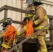 Three firefighters in full turnout gear are ascending a wooden staircase at what appears to be a training facility. They are passing and carrying coiled orange rescue ropes as they climb the stairs. The scene depicts a coordinated training exercise involving rope handling and stair navigation.