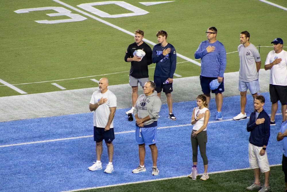 A group of people are standing on a blue-turfed football field, most with their right hands placed over their hearts, indicating they are participating in a national anthem or pledge ceremony. Several individuals are wearing Carolina Football branded apparel, suggesting this is associated with the University of North Carolina (UNC) Tar Heels football program. The scene appears to take place during a pre-game or special event on the field.