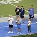 A group of people are standing on a blue-turfed football field, most with their right hands placed over their hearts, indicating they are participating in a national anthem or pledge ceremony. Several individuals are wearing Carolina Football branded apparel, suggesting this is associated with the University of North Carolina (UNC) Tar Heels football program. The scene appears to take place during a pre-game or special event on the field.