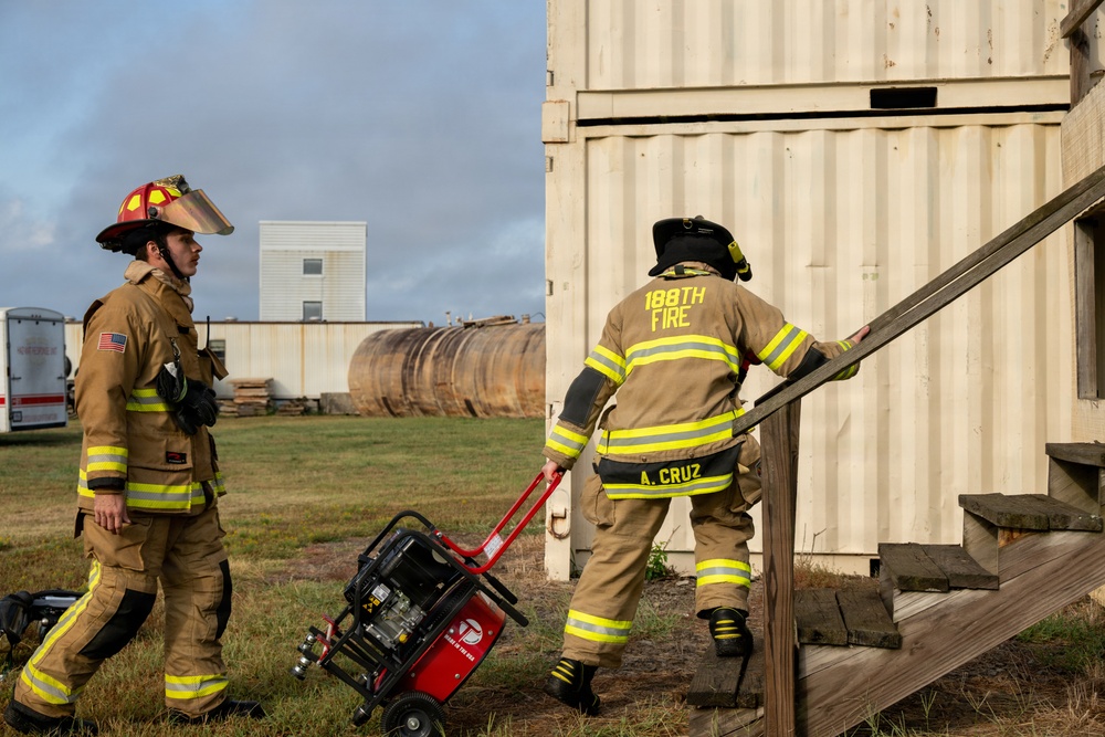 Two firefighters in full turnout gear are working near a corrugated metal structure that appears to be a training prop or shipping container. One firefighter, identified as 'A. Cruz' from the '188th Fire' unit, is climbing wooden stairs toward the structure, while the other is pulling a red portable generator. The setting appears to be an outdoor training facility with industrial equipment visible in the background.