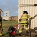 Two firefighters in full turnout gear are working near a corrugated metal structure that appears to be a training prop or shipping container. One firefighter, identified as 'A. Cruz' from the '188th Fire' unit, is climbing wooden stairs toward the structure, while the other is pulling a red portable generator. The setting appears to be an outdoor training facility with industrial equipment visible in the background.