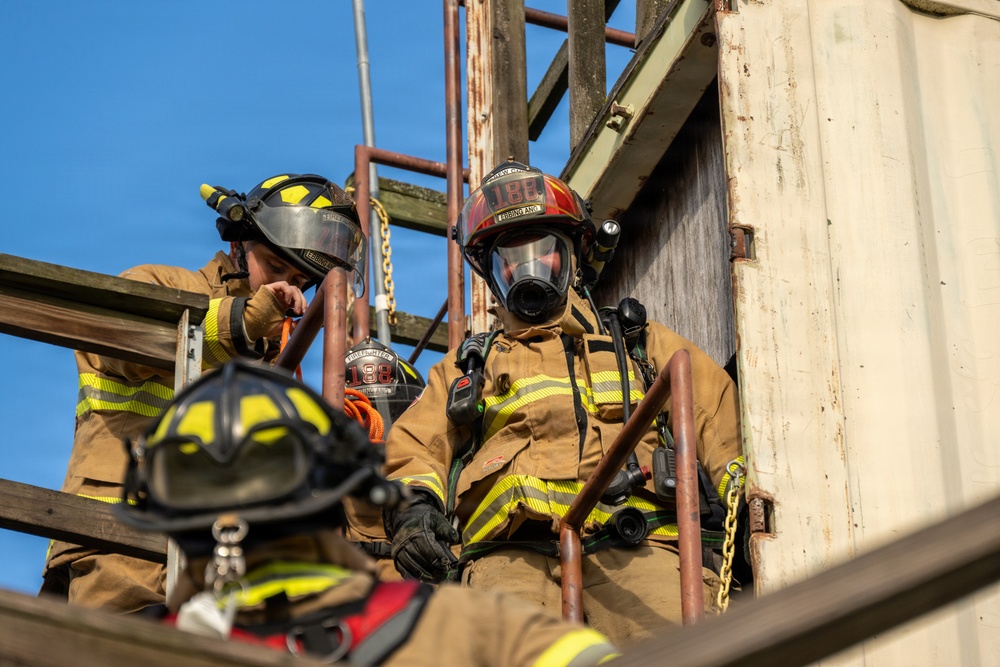 The image shows multiple firefighters in full turnout gear on what appears to be a training structure or elevated platform. At least three firefighters are visible, equipped with helmets, SCBA masks, radios, and protective clothing. The scene appears to be a firefighting training exercise or active operational response.
