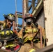 The image shows multiple firefighters in full turnout gear on what appears to be a training structure or elevated platform. At least three firefighters are visible, equipped with helmets, SCBA masks, radios, and protective clothing. The scene appears to be a firefighting training exercise or active operational response.